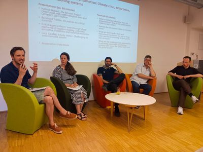 Ein Panel mit fünf Personen sitzt vor einem runden weißen Tisch. Dahinter ist eine von einem Beamer projizierte Präsentation.. Ein Panel mit fünf Personen sitzt vor einem runden weißen Tisch. Dahinter ist eine von einem Beamer projizierte Präsentation..