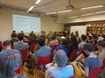 Ein Publikum sitzt in einem Saal und schaut auf ein Panel mit ein paar Personen. Ein Publikum sitzt in einem Saal und schaut auf ein Panel mit ein paar Personen.
